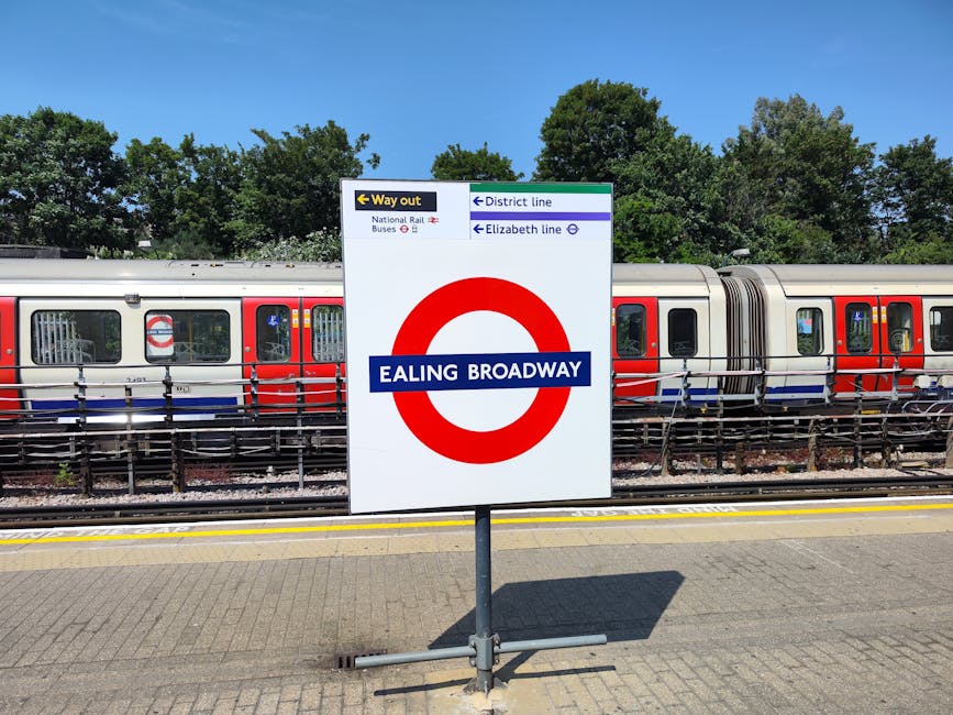 A photograph taken at an outdoor train station platform featuring a prominent sign indicating 'Ealing Broadway' in blue and white with a red circular symbol. Behind the sign, a modern train with a sil