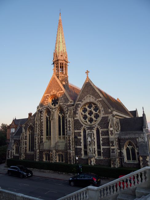 A paved pathway leading through a park area on a clear day, with leafless trees on either side casting shadows on the ground. In the background, residential houses and a church steeple are visible, wi