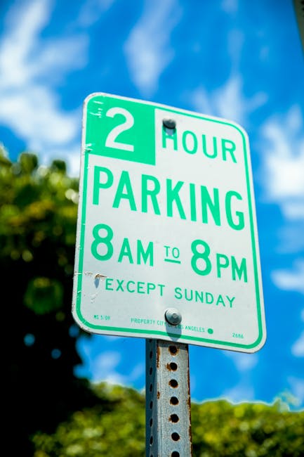 A close-up image of a green and white parking restriction sign mounted on a metal pole, indicating a two-hour parking limit from 8 am to 8 pm, excluding Sundays, in the vicinity of Northfields Station, Ealing. The sign is outdoors with a bright blue sky and white clouds visible overhead, alongside blurred green foliage in the background. It appears to be taken during daylight hours. The sign's specifications include details such as parking hours, exceptions, and local authority markings, relevant to parking regulations near residential or commercial areas. The sign may be associated with house removals or relocation services, such as those provided by Movers Ealing, assisting with parking compliance during home relocation or furniture transport within the Northfields area.