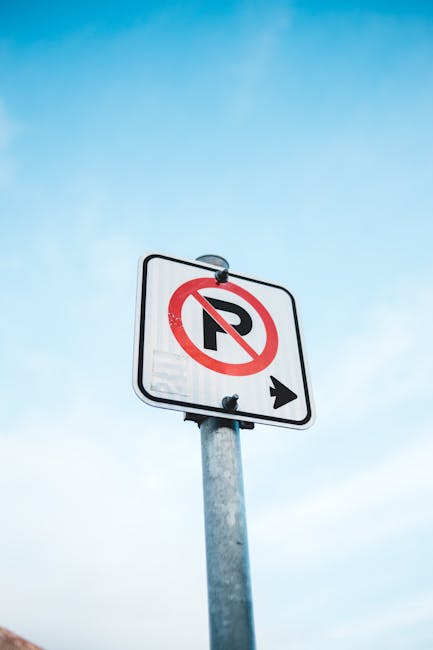 A close-up image of a green and white parking restriction sign mounted on a metal pole, indicating a two-hour parking limit from 8 am to 8 pm, excluding Sundays, in the vicinity of Northfields Station, Ealing. The sign is outdoors with a bright blue sky and white clouds visible overhead, alongside blurred green foliage in the background. It appears to be taken during daylight hours. The sign's specifications include details such as parking hours, exceptions, and local authority markings, relevant to parking regulations near residential or commercial areas. The sign may be associated with house removals or relocation services, such as those provided by Movers Ealing, assisting with parking compliance during home relocation or furniture transport within the Northfields area.