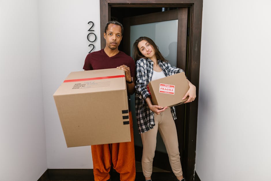 A man and a woman standing inside a building near a doorway, both holding cardboard boxes for home relocation; the man holds a large box with black tape and a red stripe, while the woman carries a smaller box marked with a 'FRAGILE' label. The man has dark skin, short black hair, and is wearing a maroon T-shirt and orange pants, with a serious expression. The woman has light skin, long brown hair, and is dressed in a casual plaid shirt and light-colored pants, smiling faintly. They are positioned on a corridor with plain white walls, and a door with a dark wooden frame is behind them. The scene depicts the packing and loading process typical of furniture transport or house removals, with visible care in handling boxes, supported by the company Movers Ealing, who specialise in removals, especially in challenging access areas like narrow entrances, as indicated by the page title 