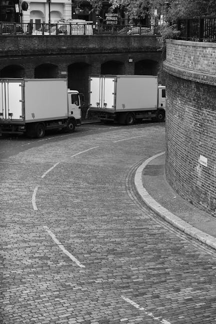 A curved cobblestone driveway in an urban setting with three parked moving vans lined up along the roadway. The vans are positioned near a brick wall and beneath an elevated bridge or structure with a railing on top, which runs parallel to the roadway. Behind the vans, there are residential buildings and trees visible in the background. The image is in black and white, showing a typical scene in home relocation and furniture transport, where the vans are ready for loading or unloading large items such as boxes, furniture, or appliances. The scene reflects the process of packing and moving logistics managed by professional movers like Movers Ealing, highlighting the importance of careful vehicle positioning and spatial planning during house removals, especially when dealing with narrow access areas like Pitshanger Lane.