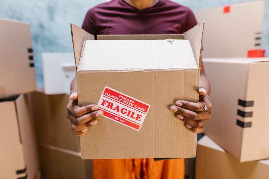 A man and a woman standing inside a building near a doorway, both holding cardboard boxes for home relocation; the man holds a large box with black tape and a red stripe, while the woman carries a smaller box marked with a 'FRAGILE' label. The man has dark skin, short black hair, and is wearing a maroon T-shirt and orange pants, with a serious expression. The woman has light skin, long brown hair, and is dressed in a casual plaid shirt and light-colored pants, smiling faintly. They are positioned on a corridor with plain white walls, and a door with a dark wooden frame is behind them. The scene depicts the packing and loading process typical of furniture transport or house removals, with visible care in handling boxes, supported by the company Movers Ealing, who specialise in removals, especially in challenging access areas like narrow entrances, as indicated by the page title 